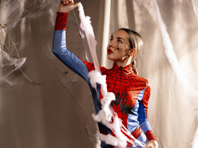 LettieSweet with a Spider-Man costume, red and blue, has white webbing on her arm, black star makeup on her cheek, and a confident smile, standing against a beige backdrop.