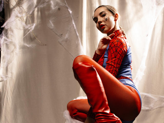 LettieSweet in a red Spider-Man suit, crouching with her ass up, wearing red fishnet stockings and red boots, looking over her shoulder with a sexy, confident expression.
