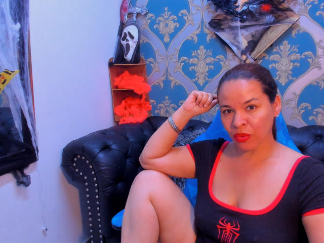 LizethBells with dark hair in a ponytail, red lipstick, black shirt with red trim, and small boobs, sitting on a black tufted couch, touching her ear.