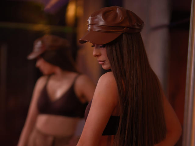 AuroraDuval with long brown hair, brown leather cap, black bra, and tan pants, standing in a dimly lit room, reflected in a mirror, showing her tits.