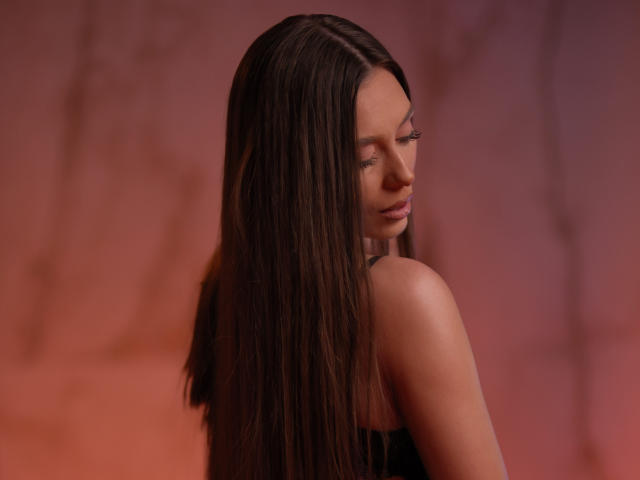 AuroraDuval with long brown hair, bare shoulders, and a serious expression, looking over her shoulder, against a warm pink and orange background. Her eyes are slightly closed.