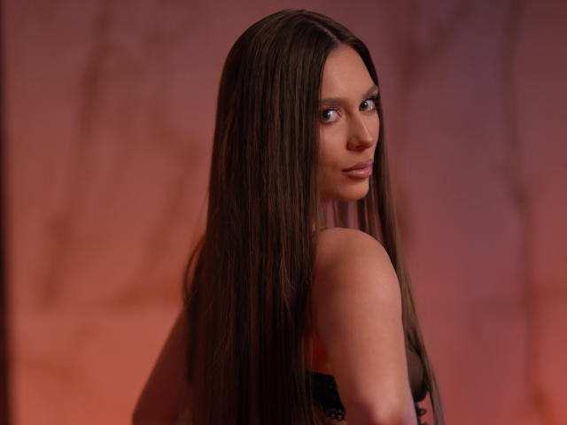 AuroraDuval with long brown hair, looking over her shoulder, showing her bare shoulders and black top, with a subtle smile, in a warm, reddish light.