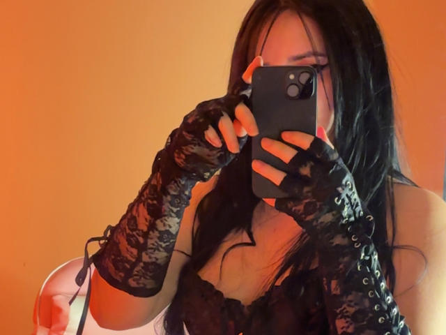 Kkatalina with long black hair, wearing black lace gloves and a lace top, taking a selfie with a black iPhone, covering her face. Her tits are slightly visible.