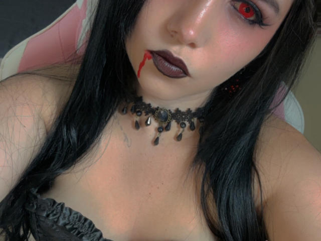 Kkatalina with black hair, red eyes, dark lipstick, and a blood drip from her mouth. She's wearing a black choker with beads and a strapless black top.