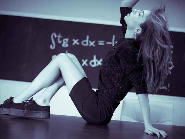 PoxyVibe with long, wavy hair, wearing a sparkly black top and black shorts, sits on a table, legs crossed, head tilted back, arms raised, in black-and-white.