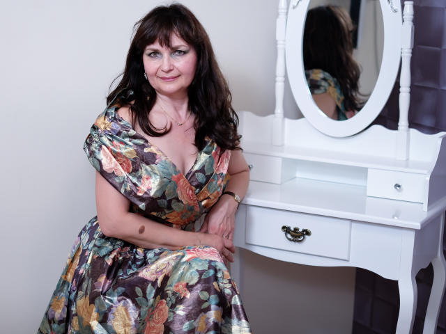 AngieGreen with brown hair, floral dress, and medium tits, sits by a white vanity with oval mirror, smiling softly, arms resting on her knees.
