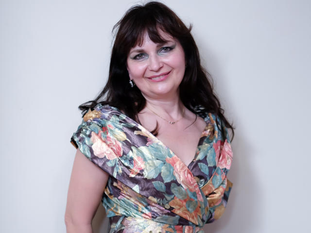 AngieGreen with dark brown hair, smiling, wearing a floral dress, exposing her small tits, standing against a plain white wall, has blue eyeshadow and a subtle gold necklace.