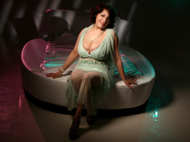 SophiaLaur, con grandes senos, sentada en una cama redonda, vestida con un vestido verde, un collar de perlas y zapatos rojos, sonriendo, con las manos sobre la cama, iluminación suave, sombras..