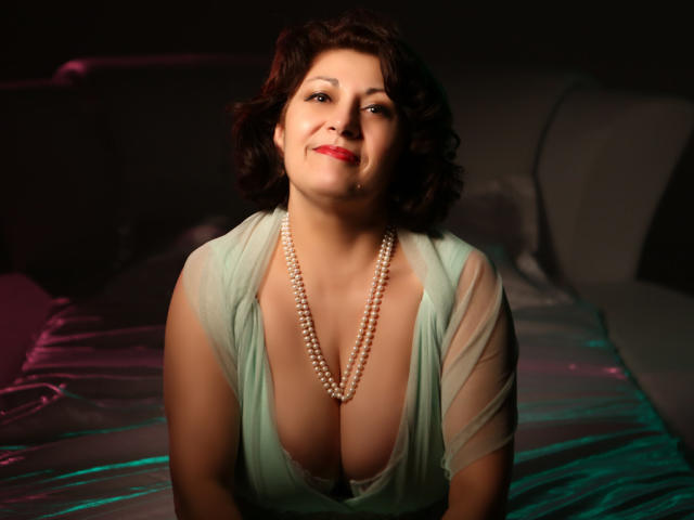 SophiaLaur, com cabelos castanhos cacheados, batom vermelho e um colar de pérolas, vestindo um vestido verde transparente, mostrando seios grandes, sentada em uma cama com lençóis verdes e rosa..