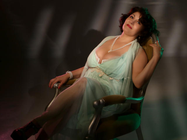 SophiaLaur, con sobrepeso y grandes senos, vestida con un vestido verde y un collar de perlas, está sentada en una silla, sosteniendo un abanico verde, mirando hacia arriba con una ligera sonrisa, cabello rizado oscuro..