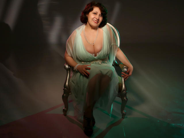 SophiaLaur, chubby with big tits, sits on a chair wearing a green dress, pearl necklace, and black heels, holding a glass, smiling with short brown hair.
