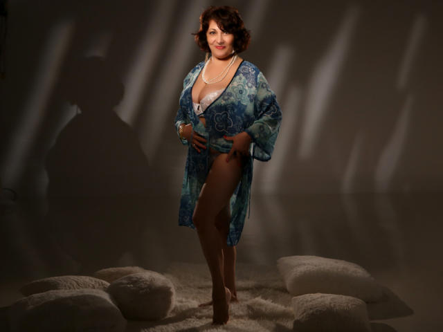 SophiaLaur has curly brown hair, blue patterned robe, white lace bra, and brown stockings, standing with one leg up, showing her tits and ass, against a shadowed backdrop.