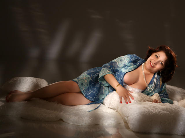 SophiaLaur, com seios grandes, deitada em pele branca, vestindo um robe azul estampado, colar de pérolas, unhas vermelhas, sorrindo, cabelo cacheado escuro, sexy, iluminação suave, em uma superfície refletora..