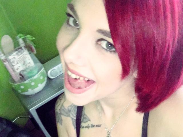 NinaDevil, con un caschetto rosso, sorride mostrando la lingua, ostentando il seno e i tatuaggi, indossando bretelle nere e una collana d'argento. I suoi occhi sono blu ed è carina..