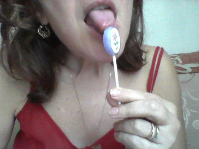 La langue de Ceryane est dehors, elle lèche un bonbon bleu et blanc avec des points de couleur. Elle porte un haut rouge à bretelles fines, qui dévoile ses seins, et elle porte une bague..