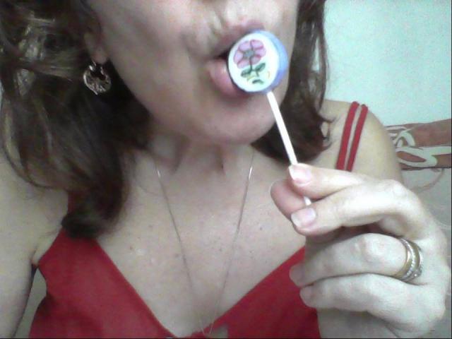 Ceryane with brown wavy hair, wearing a red bra, sucking on a lollipop with a pink flower design, holding it with her right hand, showing a gold ring.