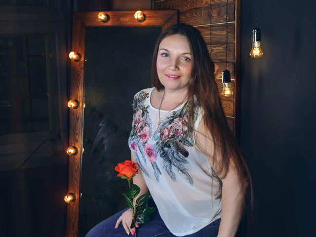 RysaLocka with long brown hair, floral shirt, holding red rose, standing by mirror with bulbs, smiling, dark background, pink lipstick, blue eyes, cute, sexy, attractive.