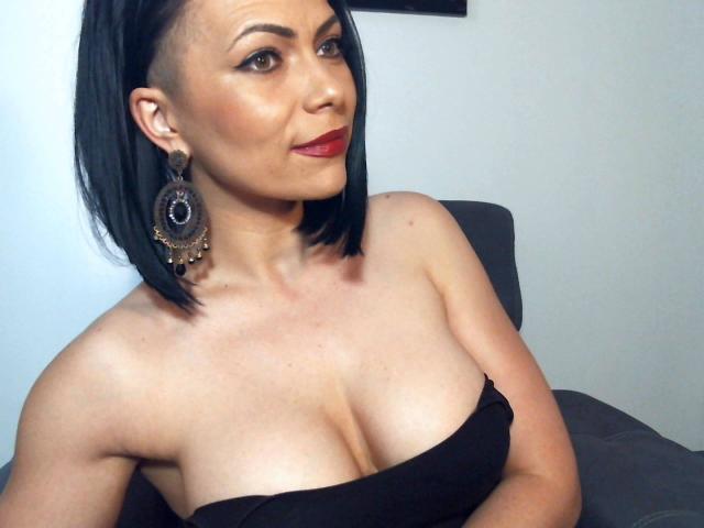 DeeLicious hat schwarzes, schulterlanges Haar, roten Lippenstift und große Brüste und trägt ein schwarzes Top ohne Träger. Sie trägt große, aufwendige Ohrringe und sitzt auf einem grauen Stuhl..