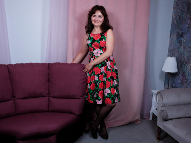 AngieGreen, with medium tits, wearing a floral dress, black stockings, and black heels, stands by a pink couch, smiling, in a room with pink curtains and a white lamp.