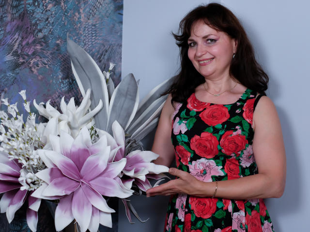 AngieGreen with medium brown hair, pink eyeshadow, floral dress, pointing at large purple-and-white lilies, smiling. She's holding her tits gently with her right hand.