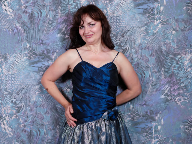AngieGreen, avec une poitrine de taille moyenne, portant un haut en satin bleu et une jupe argentée brillante, les mains sur les hanches, souriante, cheveux bruns, debout devant un mur bleu-gris texturé..