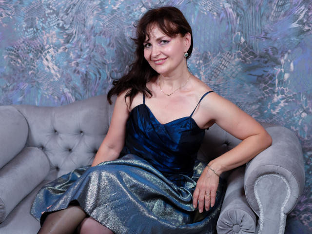 AngieGreen with medium tits, wearing a blue velvet dress and shiny silver skirt, sits on a gray tufted couch, smiling, brown hair, blue eyes, light skin, ring on right hand.