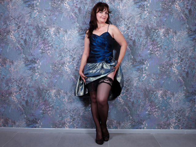 AngieGreen mit dunkelbraunen Haaren, die ein blaues Kleid trägt und es anhebt, um schwarze Strumpfhosen und ein Strumpfband zu zeigen, steht auf schwarzen High Heels und lächelt in die Kamera..