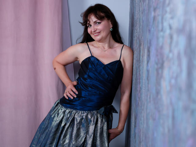 AngieGreen, con un busto de tamaño medio, vestida con una blusa de satén azul y una falda plateada brillante, de pie con una mano en la cadera, sonriendo, cabello castaño, aretes azules, cortina rosa a la izquierda..
