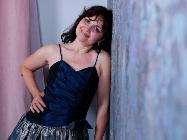 AngieGreen, avec ses cheveux brun foncé, souriante, portant un haut en satin bleu marine et une jupe argentée ornée de sequins, appuyée contre un mur texturé, une main sur la hanche, montrant sa petite poitrine..