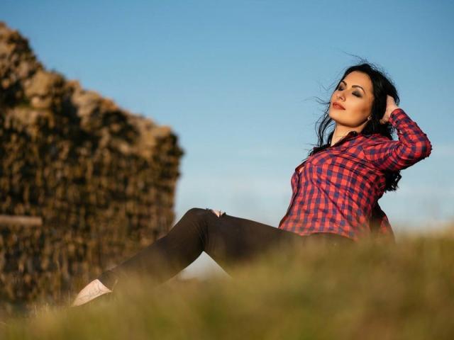 XCarla with long black hair, red plaid shirt, black pants, and red lipstick, sitting on grass, hand in hair, legs apart, blue sky, stone wall in background.