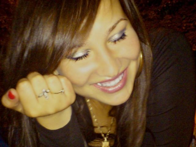 SexyJenniferOne com cabelo castanho, sorrindo, olhos fechados, vestindo uma blusa preta, colar de ouro, anéis, unha vermelha, levantando o punho, mostrando dentes brancos, lábios brilhantes e rosto fofo..