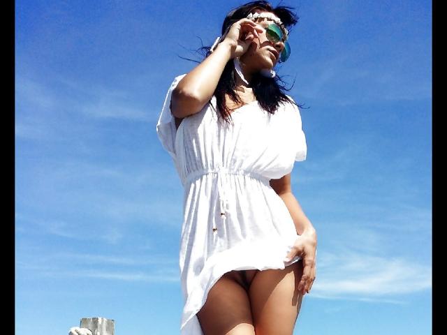 CassandraMichelli in a white dress, showing her pussy, with her hand on her head, wearing green sunglasses, against a bright blue sky. Her dress is lifted slightly.