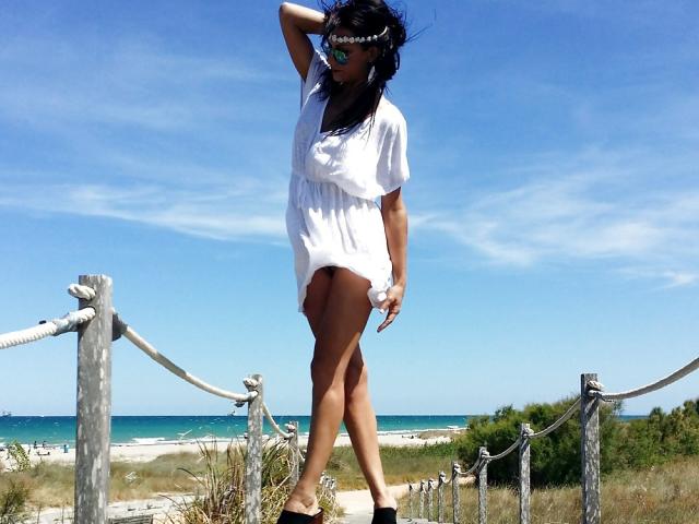CassandraMichelli stands in a white dress, black heels, and sunglasses, with one hand in her hair, showing her ass and a fluffy white wrist accessory, on a sunny beach.