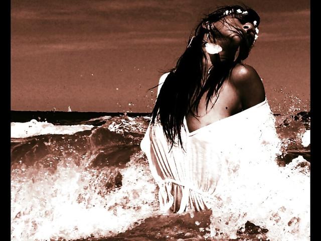CassandraMichelli with wet, long dark hair, eyes closed, wearing a white, off-shoulder dress, standing in ocean waves, dress clinging to her tits, waves splashing around her.