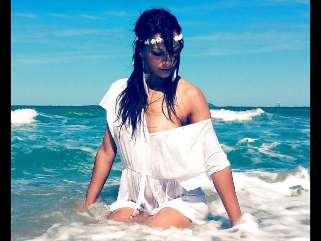 CassandraMichelli with wet black hair, white dress slipping off her boobs, flower headpiece, kneeling in ocean waves, blue sky, and white foam, looking down, arms in water.