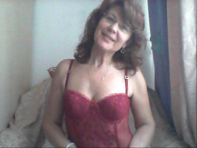 Ceryane, avec ses cheveux bruns bouclés, souriante, portant une combinaison en dentelle rouge, montrant ses seins, se tient dans une pièce aux murs clairs et avec un rideau..