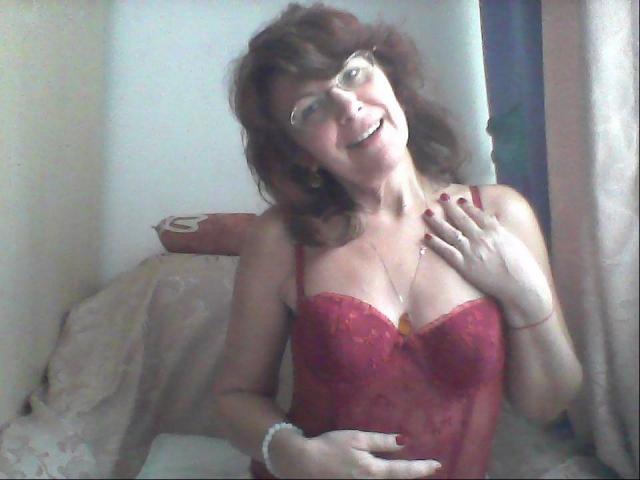Ceryane, com cabelo castanho médio, óculos e lingerie vermelha de renda, sorrindo, com a mão no peito, mostrando os seios, sentada em uma cama com um travesseiro rosa..