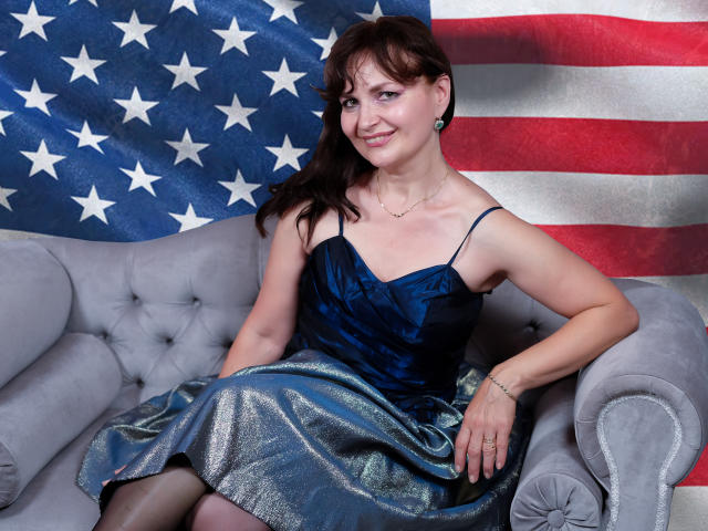 AngieGreen with brown hair, wearing a blue satin dress and shiny silver skirt, sitting on a gray couch, smiling, showing her small tits, against a large American flag.