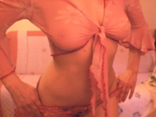 Venusya wearing a pink, see-through, tied-up top exposing her big boobs, with hands on her pink, floral-patterned panties, in a dimly lit, pink-themed room.
