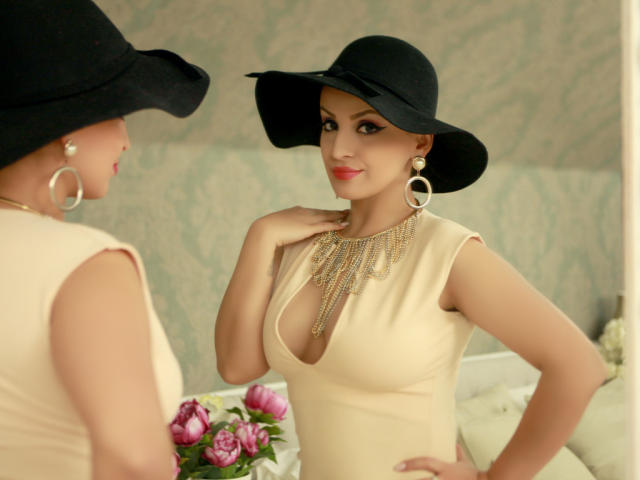 DashingFoxyX with tits in a beige dress, black hat, and gold necklace, wearing large hoop earrings, looking in a mirror, smiling, holding her chest. Roses in the background.
