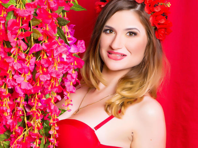 SweetLipsJenny with red bra, pink and red flowers, and red hair, smiling with lipstick. Her tits are small to medium, and she has a cute mole on her right side.