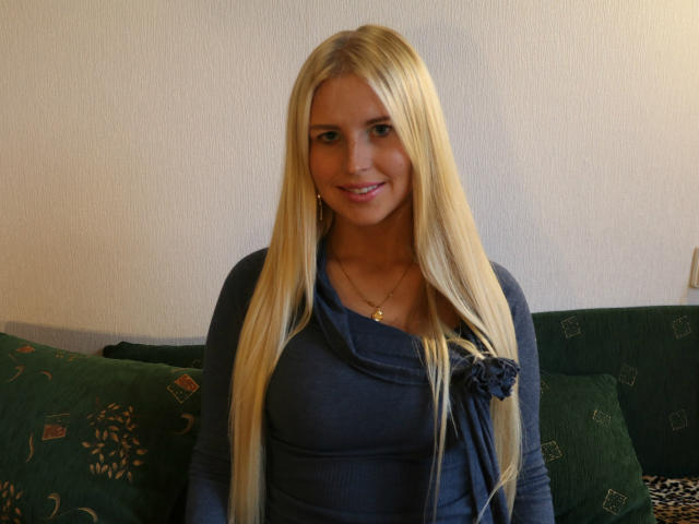 VittaHot69 com cabelo loiro comprido, blusa azul, pequena rosa no lado esquerdo, sorrindo para a câmera, usando colar de ouro, almofadas com padrão verde ao fundo, sentada em um sofá verde..
