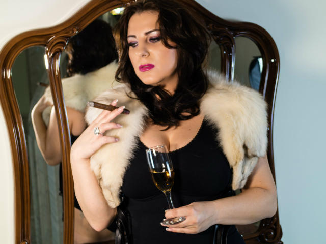 VelvetManttis with curly brown hair, pink lipstick, black dress, fur vest, holding champagne glass, reflection in wooden mirror, finger on lip, looking sultry. Boobs visible, tits prominent.