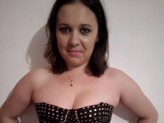 ChaudeBella69, avec des cheveux bruns moyens, porte un haut noir à pois sans bretelles, laissant entrevoir ses seins. Elle a un petit collier et un léger sourire, et se tient devant un mur blanc uni..