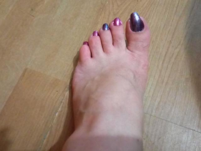 Der Fuß von KaryQueen mit pinken und lila glänzenden Zehennägeln auf einem Holzboden, wobei ein blauer Nagellack am großen Zeh und pink am zweiten Zeh zu sehen ist..