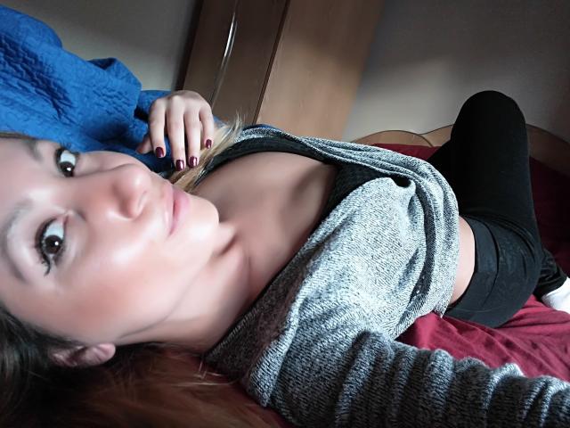 GinnyGold sdraiata su un letto, indossa un maglione grigio aperto, una maglietta nera che rivela piccoli seni, pantaloni neri, un letto rosso, una coperta blu, una mano con le unghie viola, che guarda la fotocamera..