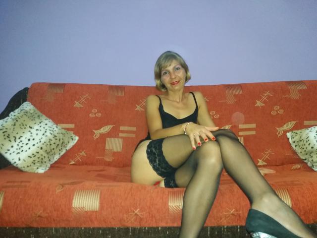 Bobandchris69, eine blonde Frau mit kurzen Haaren, trägt schwarze Spitze, hohe Strümpfe und grüne Schuhe. Sie sitzt auf einem roten Sofa, die Beine gekreuzt, trägt roten Lippenstift und ein Armband..
