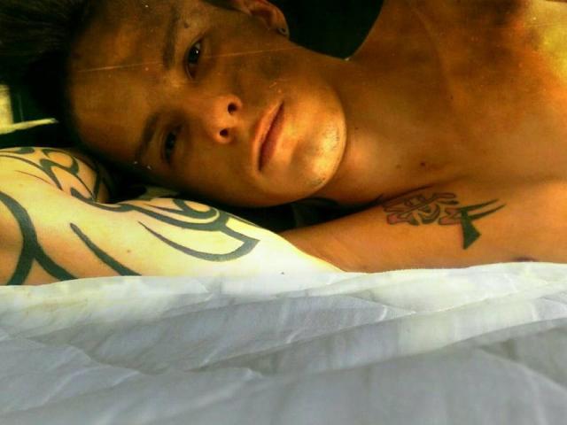 YeremyWalker, senza camicia, è sdraiato su un letto con lenzuola bianche, ha un tatuaggio sul braccio destro e sul petto, e un altro tatuaggio sul lato sinistro, sembra stanco, con il viso illuminato dal sole e i capelli castani..