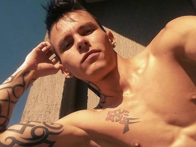 YeremyWalker, sem camisa e com tatuagens, tem cabelo curto e escuro, piercings e um peito musculoso. Ele está encostado em uma parede texturizada, olhando para a câmera com uma expressão séria..