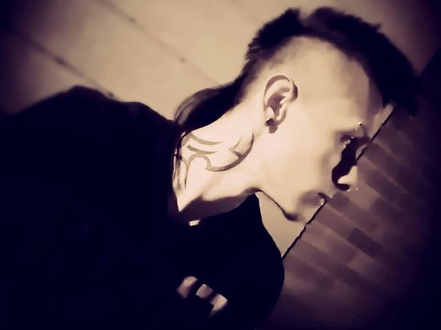 Yeremy Walker, con un peinado estilo mohawk, mirando hacia la derecha, tiene un tatuaje tribal en el cuello y un piercing en la nariz, y lleva una camisa negra, en una fotografía con poca luz y tonos sepia..